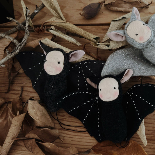 Two black bat-shaped toys with pink faces on a wooden surface with leaves.