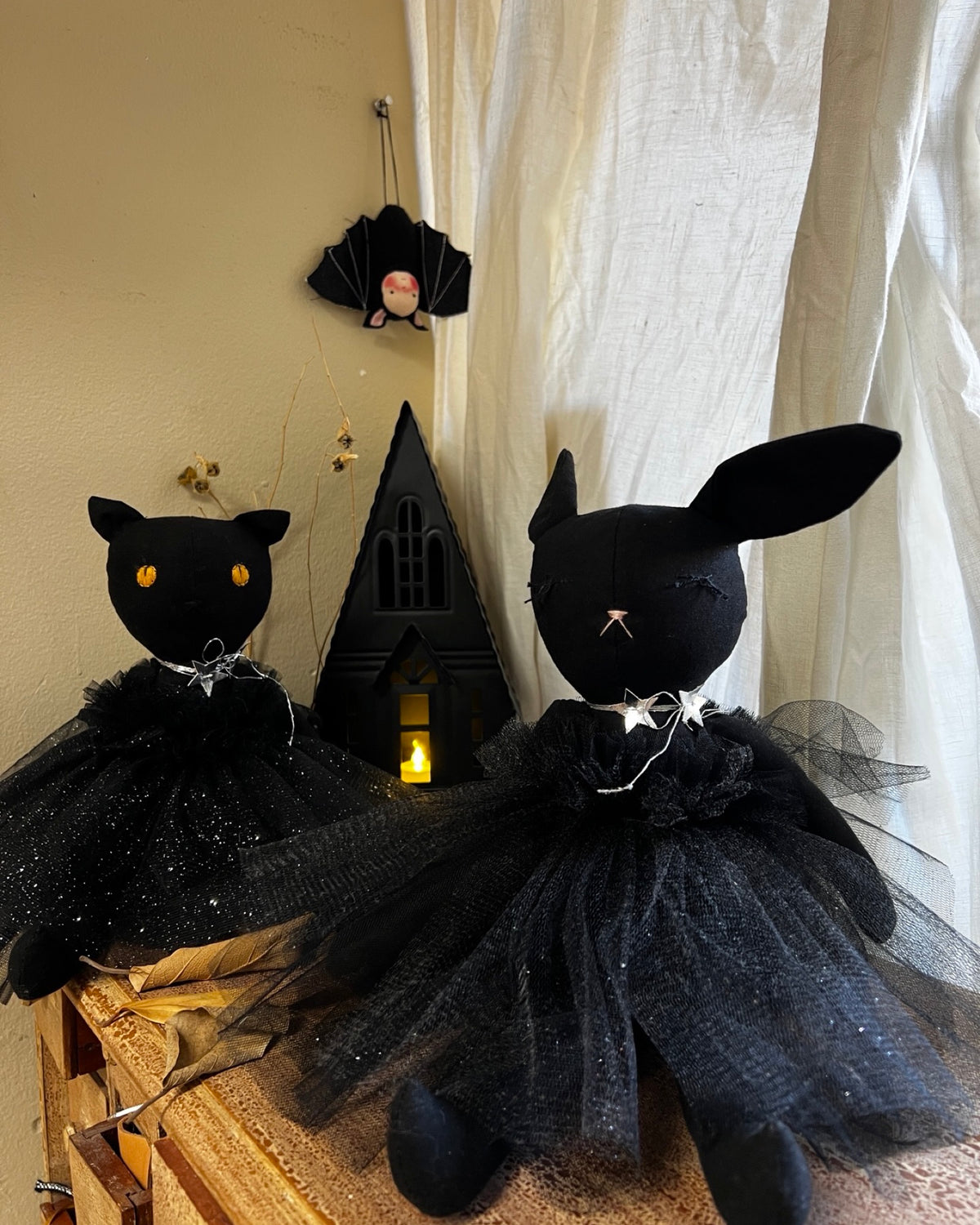 Black cat and  bunny plush dolls wearing black tutus, displayed on a wooden surface with a small black house decoration and white curtain in the background.