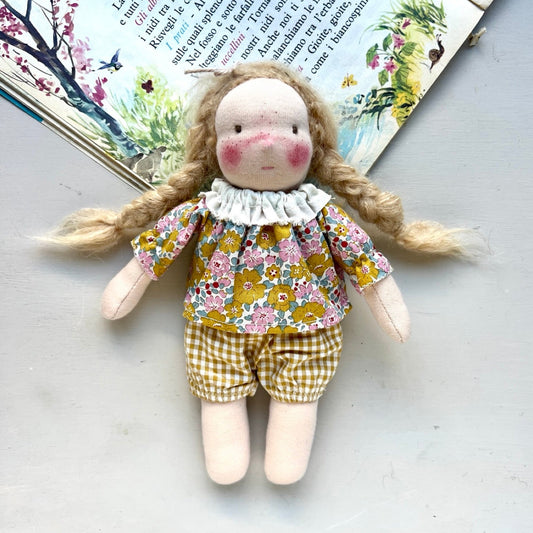 Handmade doll with floral top and checkered shorts on a light surface with a book in the background.