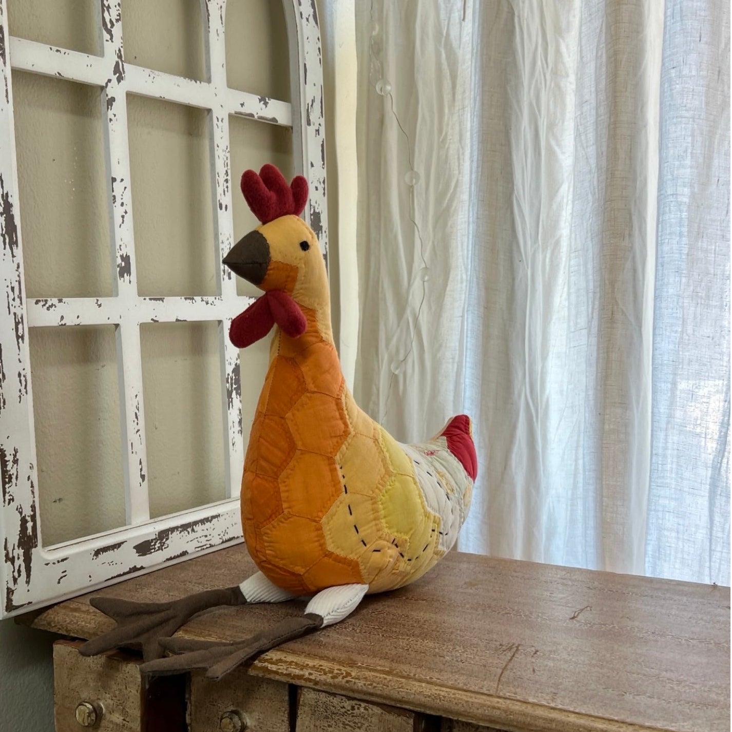 Plush rooster toy on a wooden surface with a decorative window frame in the background.